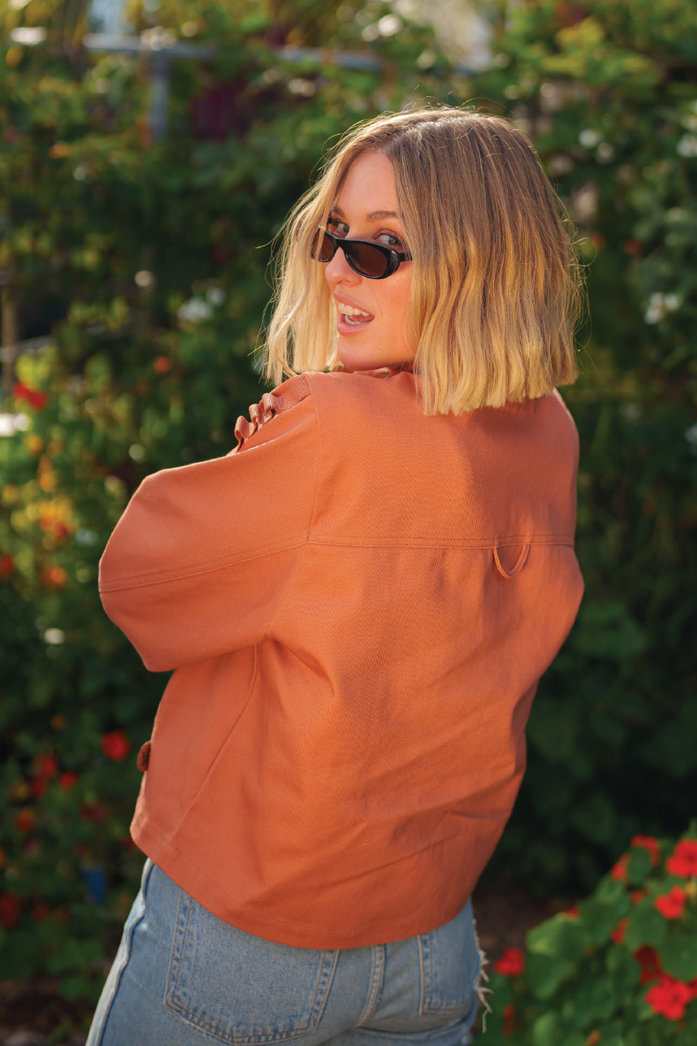 blonde woman smiling while looking over shoulder wearing oval sunglasses coral red denim 4-pocket long sleeve outdoor denim jacket and Levis jeans with garden in background