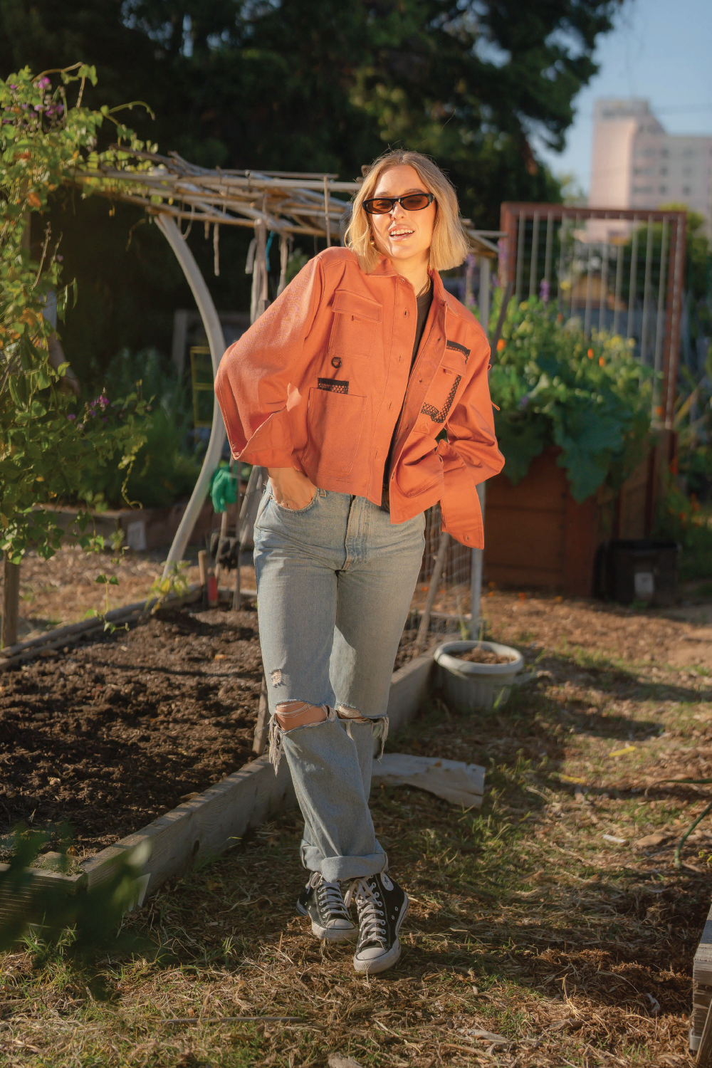 blonde woman with short hair smiling wearing coral red denim 4-pocket long sleeve outdoor denim jacket and Levis jeans and black converse with garden in background