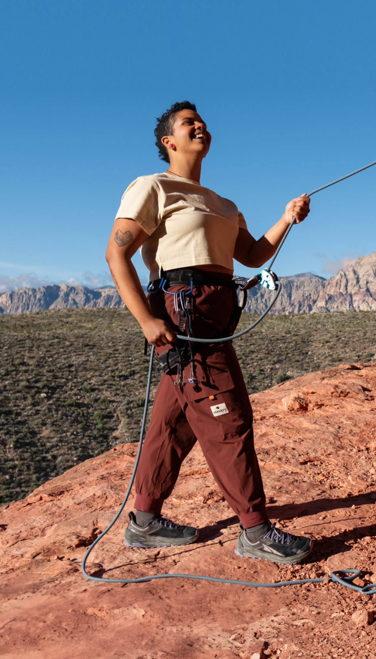 Tall woman with short hair, sand colored crop top and climbing pants preparing to climb a red rock with her climbing gear