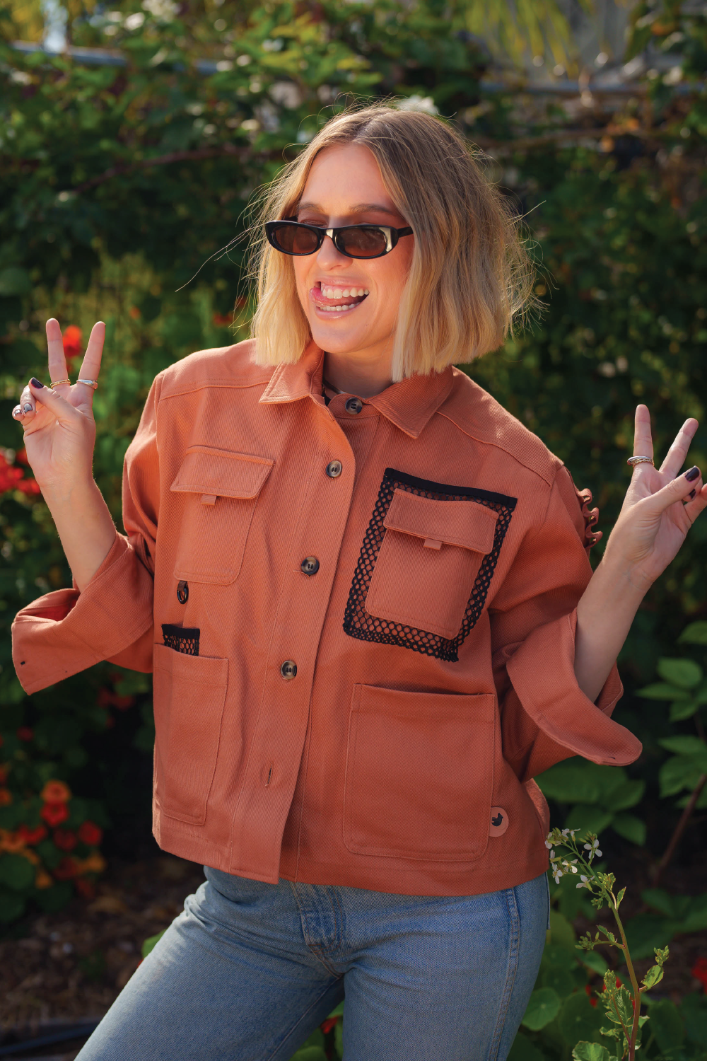 blonde woman with short hair smiling wearing coral red denim 4-pocket long sleeve outdoor denim jacket and Levis jeans with garden in background