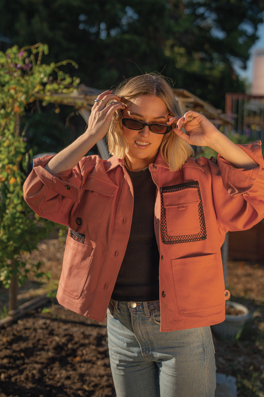 blonde woman with short hair smiling wearing oval sunglasses coral red denim 4-pocket long sleeve outdoor denim jacket and Levis jeans and black top with garden in background