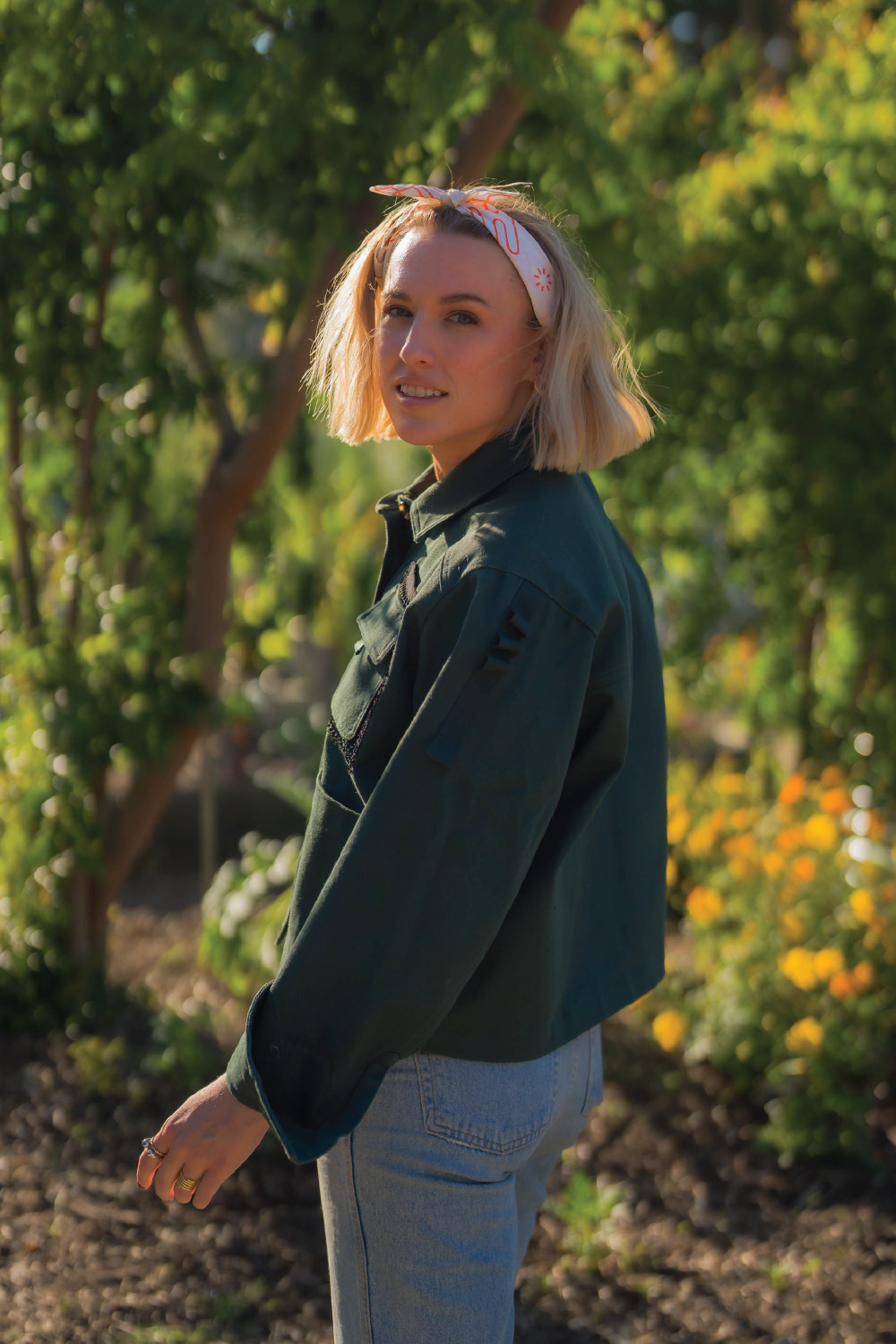 Blonde woman looking over shoulder wearing forest green long sleeve denim jacket with sleeve detail front pockets denim Levis jeans and head scarf and black converse originals