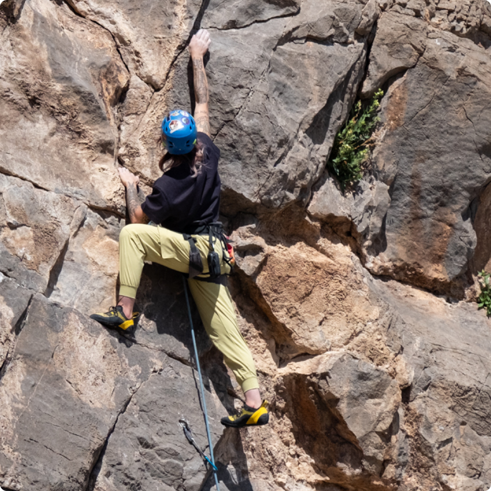 women in yellow green climbing pants climbing a mountain rock with climbing gear and helmet