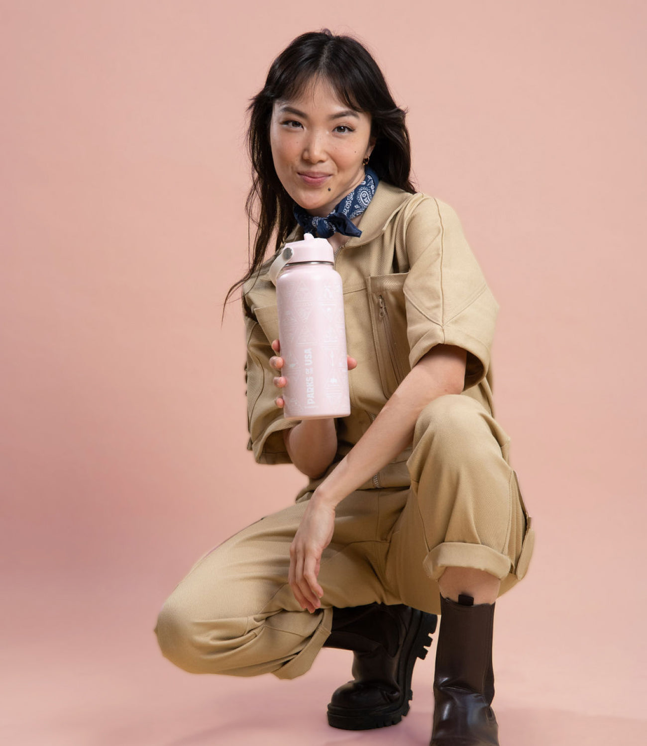 asian woman with brown hair and brown wearing a khaki denim cargo jumpsuit and boots while holding pink water travel bottle