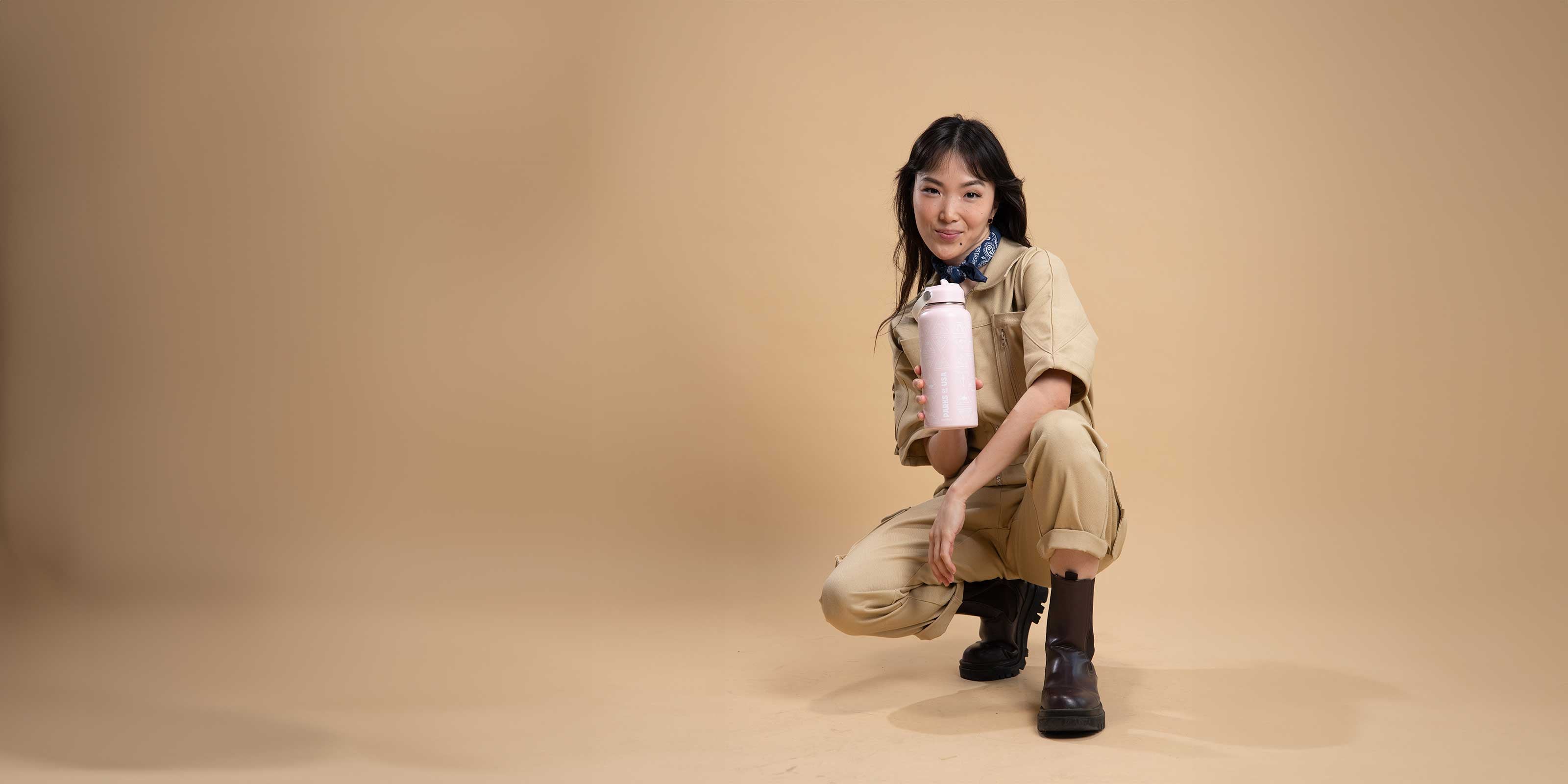 asian woman crowching down wearing khaki denim jumpsuit with boots and holding pink travel water bottle