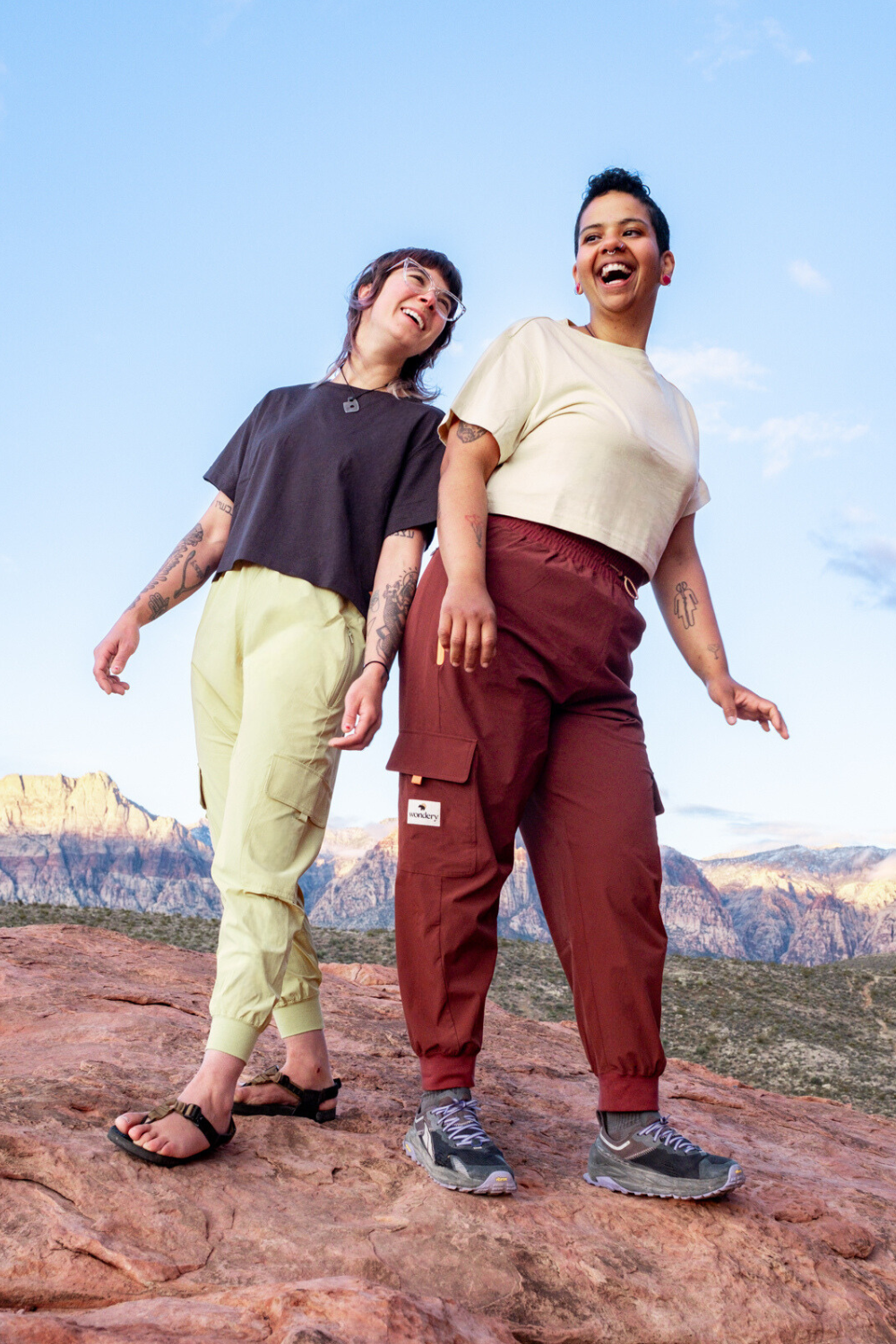 two women of different height smiling on top of red rocky mountain with crop tops and cargo pocket lightweight climbing pants with chaco sandals and sneeakers