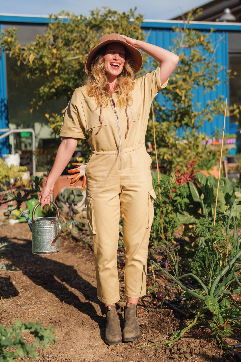 tall blonde woman in wide brim summer hat and short sleeve jumpsuit with boots and watering can in Los Angeles community garden
