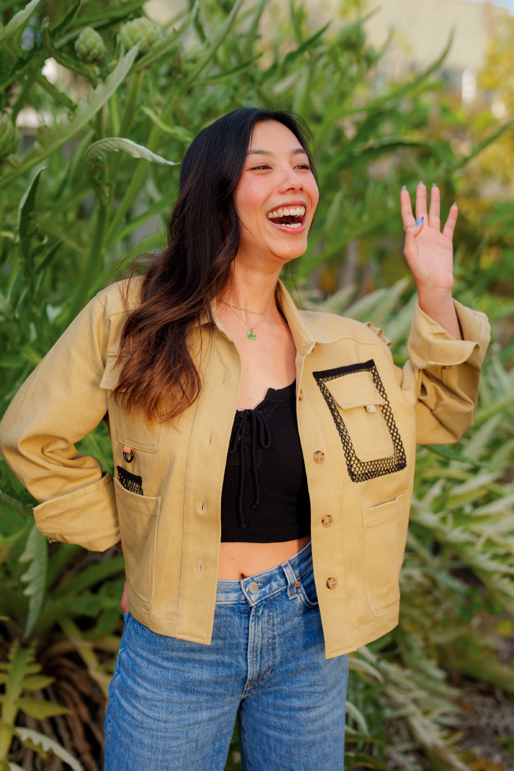 asian woman smiling while in khaki brown denim jacket covering black crop top with long sleeves and denim Levis jeans with garden in background