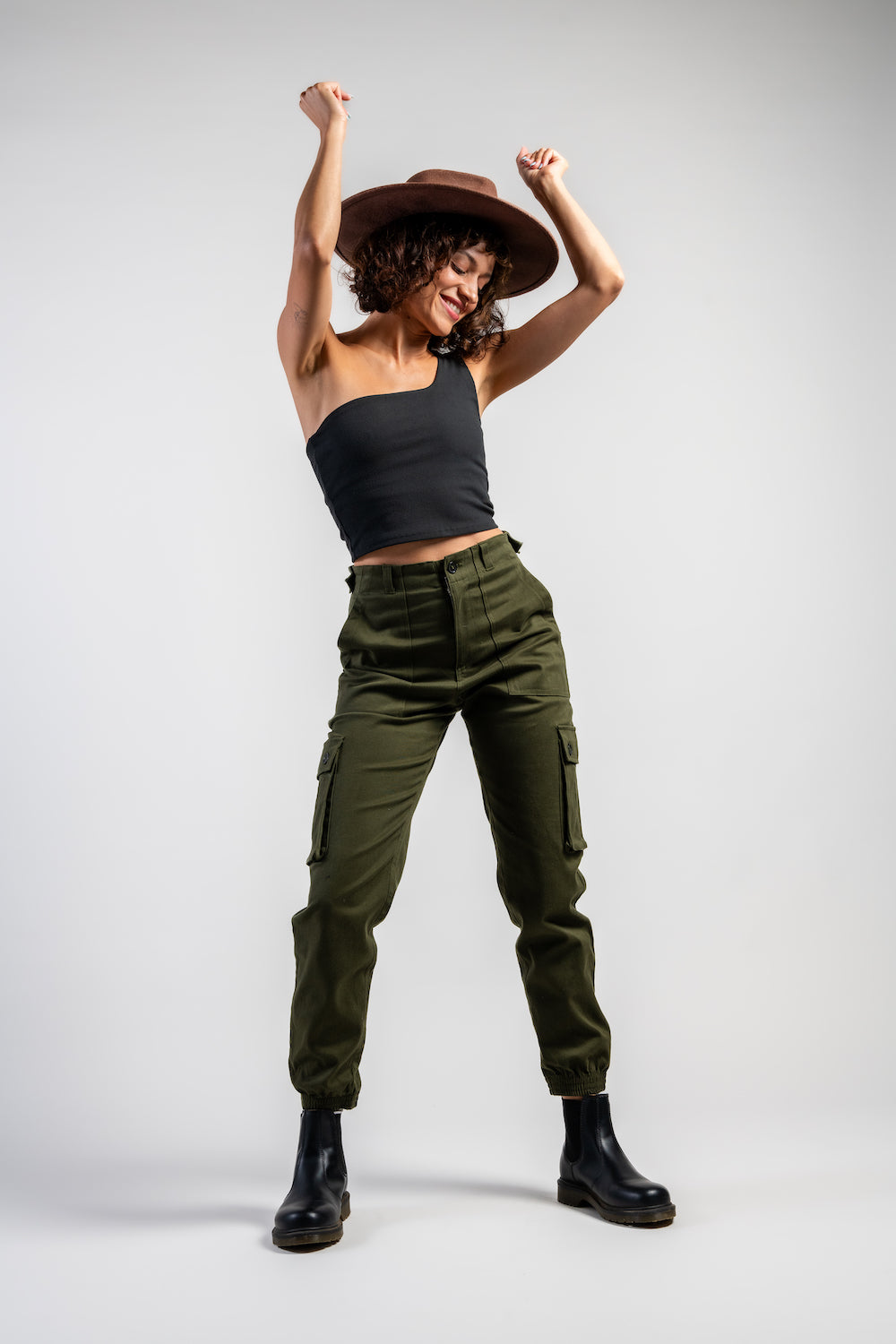 woman in army green cargo outdoor hiking camping pants and asymmetrical black top and wide brim hat #color_forest